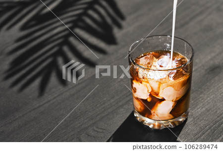 Iced coffee in a glass with cream and ice cubes on a black wooden table, selective focus, close-up. Cream is poured into a glass. Cold summer drink made from coffee and milk, cooking concept 106289474