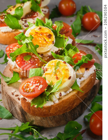 Sandwiches with soft cottage cheese or cheese, tomatoes, arugula and egg, sprinkled with sesame seeds are placed on a rustic wooden board. Healthy food, toast bruschetta, close-up selective focus Sandwiches with soft cottage cheese or cheese, tomatoes, arugula and egg, sprinkled with sesame seeds are placed on a rustic wooden board. Healthy food, toast bruschetta, close-up selective focus 106290384