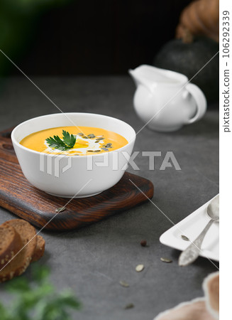 Pumpkin chowder with cream, pumpkin seeds and parsley in a white bowl on a gray background. Seasonal vegetarian soup, selective focus on parsley 106292339