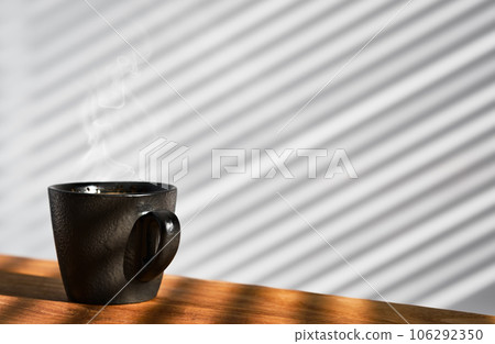 Cup of freshly made espresso coffee in a cup on a wooden table, steam rises over the ristretto. Coffee background. The wall of the cafe is covered with sunny stripes from the blinds. Coffee break Cup of freshly made espresso coffee in a cup on a wooden table, steam rises over the ristretto. Coffee background. The wall of the cafe is covered with sunny stripes from the blinds. Coffee break 106292350