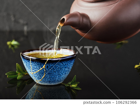 Hot green tea is poured from a ceramic teapot into a bowl. Selective focus on the blue cup. Steam will rise above the mug Reclaimed ceramic blue cup, second life of things, recycling or kintsugi 106292366