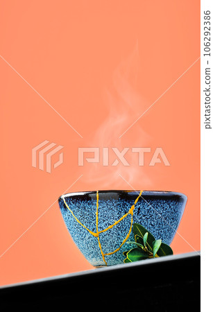 Cup of green japanese tea against a background of burnt sienna. Selective focus on cup. Steam rises above the bowl. Reclaimed ceramic blue cup, second life of things, recycling or kintsugi Cup of green japanese tea against a background of burnt sienna. Selective focus on cup. Steam rises above the bowl. Reclaimed ceramic blue cup, second life of things, recycling or kintsugi 106292386