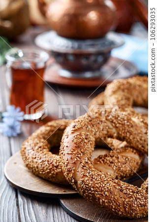 Freshly baked simit baked goods with sesame seeds close-up (Turkish bagel - Gevrek or Kuluri). Traditional white bread with sesame seeds for breakfast, a glass of armudu with red tea on a wooden table Freshly baked simit baked goods with sesame seeds close-up (Turkish bagel - Gevrek or Kuluri). Traditional white bread with sesame seeds for breakfast, a glass of armudu with red tea on a wooden table 106293035