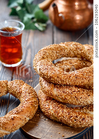 Freshly baked simit baked goods with sesame seeds close-up (Turkish bagel - Gevrek or Kuluri). Traditional white bread with sesame seeds for breakfast, a glass of armudu with red tea on a wooden table 106293040