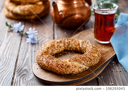 Freshly baked simit baked goods with sesame seeds close-up (Turkish bagel - Gevrek or Kuluri). Traditional white bread with sesame seeds for breakfast, a glass of armudu with red tea on a wooden table Freshly baked simit baked goods with sesame seeds close-up (Turkish bagel - Gevrek or Kuluri). Traditional white bread with sesame seeds for breakfast, a glass of armudu with red tea on a wooden table 106293043