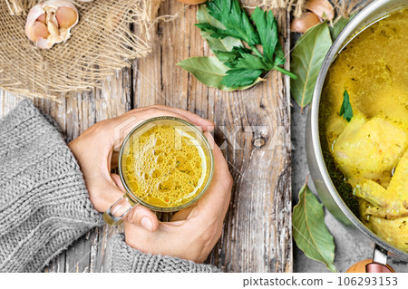 Chicken broth is poured into a glass mug, the girl's hands are holding the mug in her hands. Pan with soup and ingredients for soup on a wooden table. Flat lay 106293153