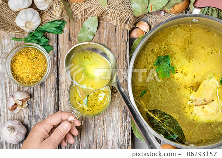 Chicken broth with vegetables and spices in a saucepan, ingredients for soup on a wooden table. Girl's hand holding a mug with broth poured from a ladle, top view 106293157