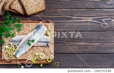 Herring fillet with parsley and onions on a cutting board, top view, dark wooden background. Traditional Norwegian or Danish smorrebrod ingredients. Herring sandwich, healthy food concept. 106293286