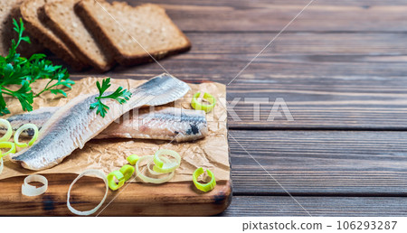 Herring fillet with parsley and onion on a cutting board, close-up, dark wooden background. Traditional Norwegian or Danish smorrebrod ingredients. Herring sandwich, healthy food concept. 106293287