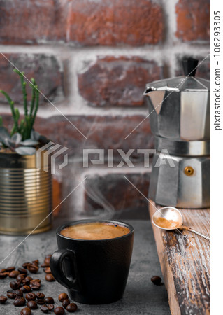 Cup of espresso coffee, steam rises above the mug, a coffee pot and coffee beans on a gray stone table, brickwork on the background. Close-up, coffee break or breakfast, coffee time 106293305