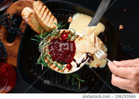 Camembert cheese in a skillet with walnuts and rosemary. Cranberry sauce with cheese. Top view, black background. Slices of baguette in a frying pan, girl's hands with cutlery over a dish 106293666