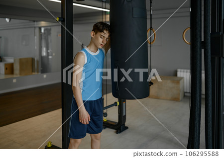 Tired boy standing nearby punch bag taking rest after hard training Tired boy standing nearby punch bag taking rest after hard training 106295588