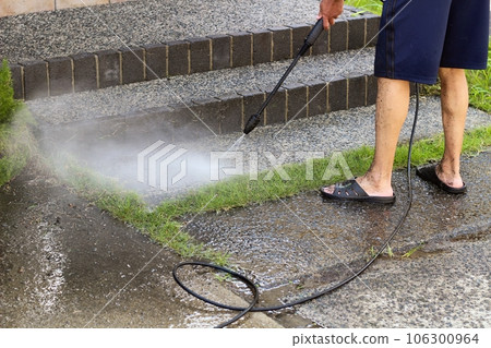 A high-pressure washer cleaning gravel-filled concrete in front of the entrance A man in shorts is working 106300964