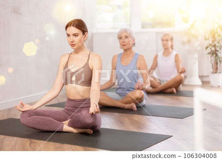 Three generations of women perform half-lotus position of Ardha Padmasana in gym 106304008