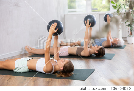 Family grandmother, mother and granddaughter perform fitness exercises lying on floor with softball. 106304030