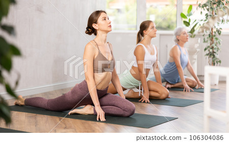 Three generations of women perform sitting pigeon pose Eka Pada Rajakapotasana in gym 106304086