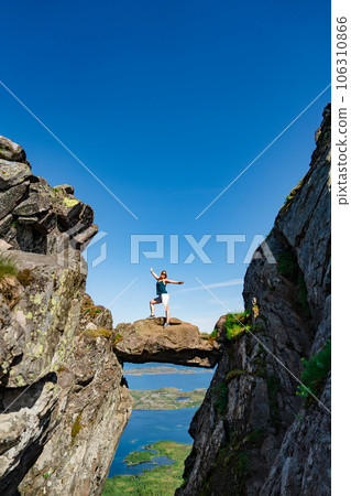 Brave traveler woman standing on hanging stone between rocks. Djevelporten in Norway Lofoten islands Brave traveler woman standing on hanging stone between rocks. Djevelporten in Norway Lofoten islands 106310866