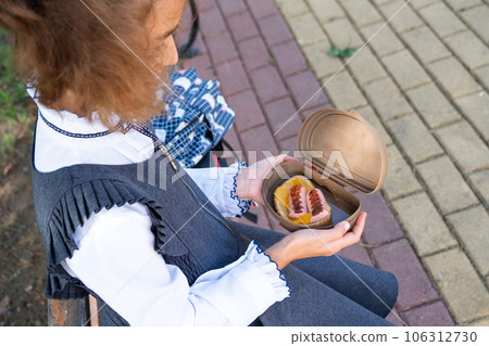 Girl with backpack eating sandwich packed in a sandwich box near school. A quick snack with a bun, unhealthy food, lunch from school. Back to school. Education, primary school classes, September 1 106312730