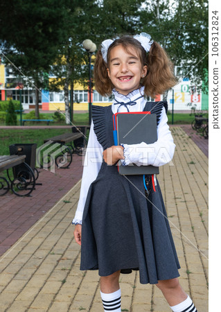 Girl with backpack, school uniform with white bows and stack of books near school. Back to school, happy pupil, heavy textbooks. Education, primary school classes, September 1 106312824
