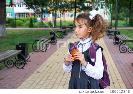 Girl with a backpack eating pie near school. A quick snack with a bun, unhealthy food, lunch from school. Back to school. Education, primary school classes, September 1 106312848