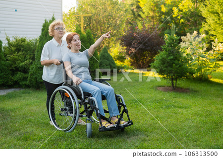 An elderly woman is carrying an adult daughter sitting in a wheelchair. Caucasian woman pointing her finger admiringly while walking outdoors. An elderly woman is carrying an adult daughter sitting in a wheelchair. Caucasian woman pointing her finger admiringly while walking outdoors. 106313500
