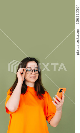 Picture of a cheerful young woman in an orange T-shirt using a modern smartphone in empty green Picture of a cheerful young woman in an orange T-shirt using a modern smartphone in empty green 106313594