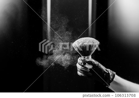 A woman shakes a large powder brush with clouds of dust on a black background. Treatment after instant tanning. Monochrome. 106313708