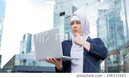 Thoughtful business woman in hijab and suit is holding a laptop outdoors. 106313941