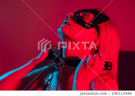 Blonde woman in leather mask putting on collar in bedroom with red blue neon light.  106313946