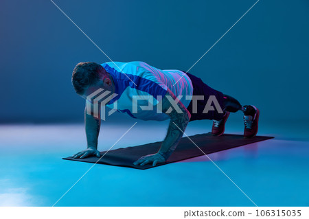 Confident man in sport uniform prosthetic legs doing push-up exercises in horizontal position in neon light on floor. Motivation poster. Confident man in sport uniform prosthetic legs doing push-up exercises in horizontal position in neon light on floor. Motivation poster. 106315035