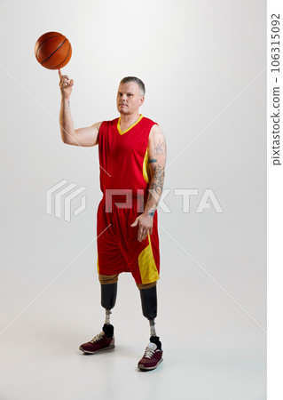 Attractive, confident, sporty man with prosthetic legs, disability in uniform spins basketball ball on finger. Inclusive sport for people with disabilities. 106315092