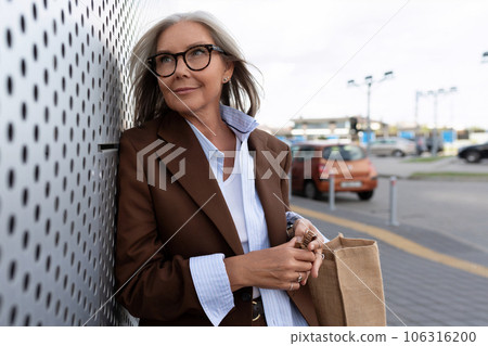 confident 60 year old gray-haired business woman dressed in a brown jacket walks around the city 106316200