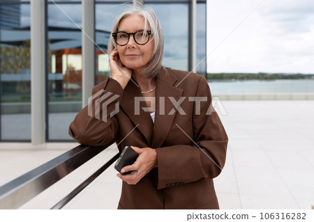 a slender senior business woman with gray hair dressed in an elegant brown jacket stands against the a slender senior business woman with gray hair dressed in an elegant brown jacket stands against the 106316282