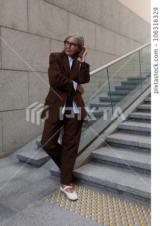 charming successful senior woman with gray hair and glasses is dressed in a brown designer suit 106316329
