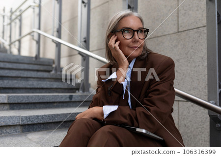 well-groomed slender mature business woman with gray hair is dressed in a brown suit in a city 106316399