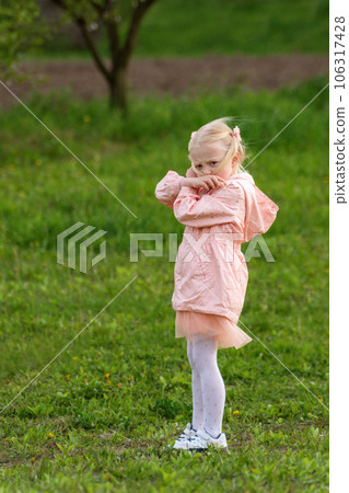 Portrait of little European blonde girl in pink jacket in green meadow or clearing. Vertical frame. 106317428