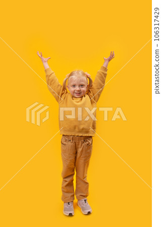Little fair-haired girl of 4-5 years old with two ponytails raised her hands up and looked into camera. Vertical yellow background 106317429