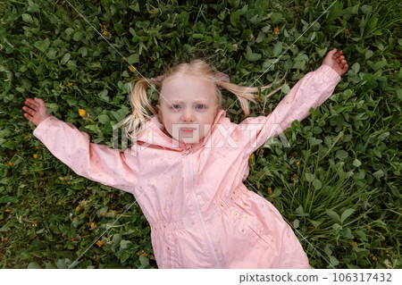 Portrait of blonde girl with two ponytails in pink jacket lies on green grass. Towheaded small girl outside. Spring day 106317432