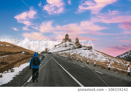 Stepantsminda, Gergeti, Georgia. Man Tourist Backpacker Traveler Photographer Walking To Holy Trinity Church - Tsminda Sameba. Beautiful Georgian Landscape In Early Winter Stepantsminda, Gergeti, Georgia. Man Tourist Backpacker Traveler Photographer Walking To Holy Trinity Church - Tsminda Sameba. Beautiful Georgian Landscape In Early Winter 106319142