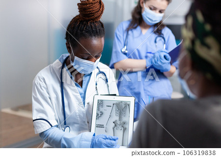 Bone doctor explaining clinical information on tablet to patient during orthopedic medical exam. Orthopedist in modern sterile hospital office wearing face mask and medical gloves 106319838