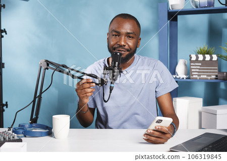 Cheerful vlogger speaking in microphone while reading from smartphone and streaming online portrait. Smiling blogger answering subscribers question and broadcasting live for social media channel 106319845