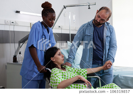 Gynecologist nurse helping pregnant woman to go to surgery in hospital ward. Patient with pregnancy sitting in wheelchair being comforting by husband and assistant in maternity facility Gynecologist nurse helping pregnant woman to go to surgery in hospital ward. Patient with pregnancy sitting in wheelchair being comforting by husband and assistant in maternity facility 106319857