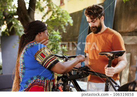 Youthful and athletic interracial couple fixing damaged bicycle outside for summer leisure cycling. Healthy black woman helping caucasian man by clamping bike frame to repair-stand for easy repairing. Youthful and athletic interracial couple fixing damaged bicycle outside for summer leisure cycling. Healthy black woman helping caucasian man by clamping bike frame to repair-stand for easy repairing. 106319858