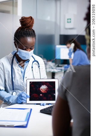 Microbiology doctor teaching patient about disease preventative protection against microscopic bacteria virus covid-19 microbe. Face mask and gloves wearing medic showing coronavirus on tablet Microbiology doctor teaching patient about disease preventative protection against microscopic bacteria virus covid-19 microbe. Face mask and gloves wearing medic showing coronavirus on tablet 106319898