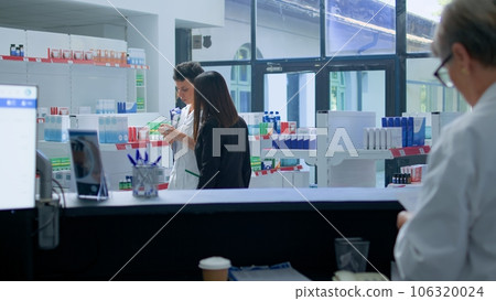 Asian client in need of prescription medicaments informations in apothecary, requesting help from cashier who redirects her to druggist checking pharmaceutical products inventory Asian client in need of prescription medicaments informations in apothecary, requesting help from cashier who redirects her to druggist checking pharmaceutical products inventory 106320024