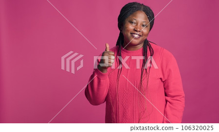 Female with african american ethnicity showing thumbs up with stunning smile on her face. Glamorous black woman confidently gesturing approval with her fingers against colorful background. 106320025