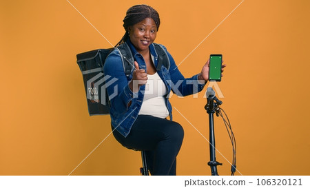 African american female with backpack carries smartphone with isolated green screen display. Friendly courier shows online ordering logistics on cell phone with chromakey mockup template. African american female with backpack carries smartphone with isolated green screen display. Friendly courier shows online ordering logistics on cell phone with chromakey mockup template. 106320121