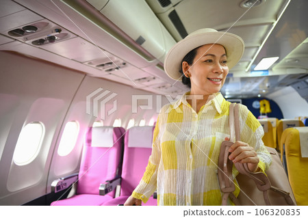 Smiling 60s female tourist with suitcase walking the aisle on plane. Travel and summer vacation concept 106320835