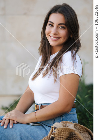 Portrait of beautiful woman in denim style sits on stone bench in front of wall. Female look into camera and smile Portrait of beautiful woman in denim style sits on stone bench in front of wall. Female look into camera and smile 106322668