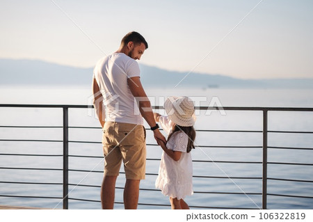 Back view of daughter with her father stand on heigh balcony in front of sea sunset. Dad with his little girl looking on mountains Back view of daughter with her father stand on heigh balcony in front of sea sunset. Dad with his little girl looking on mountains 106322819